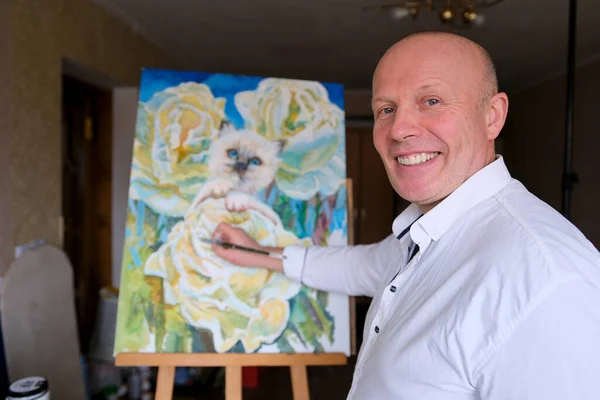 the artist man with a brush draws to a white cat with blue eyes smiles looks into frame the masters hand painting drawing flowers Bright delightful beauty on the wall decoration art
