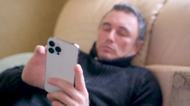 iPhone 13 Pro Max telefonunun sıradan insanlar tarafından kullanılması koyu renk ceketli bir adam kanepenin üzerinde odaktan uzak bir şekilde yatıyor ön planda beyaz telefon sosyal ağları, internet iletişimi.