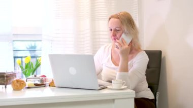 Evde dizüstü bilgisayarla çalışan küçük işletme sahibi bir kadın. Beyaz kazaklı bir kadın konuşuyor. Sarı lale koltuğu yemek masası. Yüksek kalite 4K görüntü.