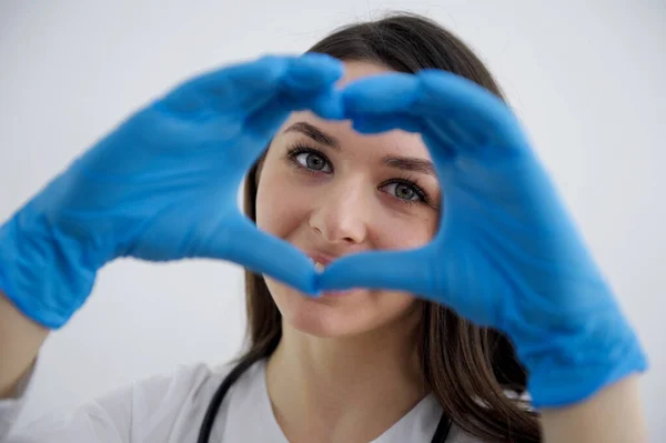 Smiling beautiful young indian ethnicity female doctor cardiologist showing heart symbol with fingers, expressing love and support to patients, healthcare medical help charity donation