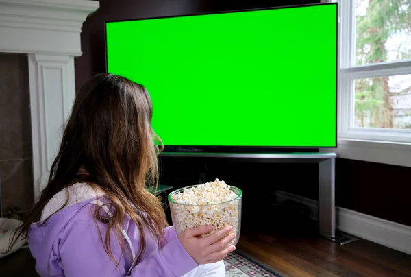young woman watches projector, TV, movies with popcorn in evening large green TV screen girl is sitting on floor near fireplace on carpet in purple jacket wearing white pants is watching movie ad