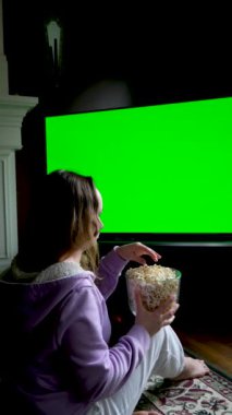 Genç kadın projektör, televizyon, akşamları patlamış mısırlı filmler büyük yeşil ekran kız, şöminenin yanında mor ceketli, beyaz pantolonlu ve beyaz pantolonlu bir şekilde oturmuş film reklamı izliyor.