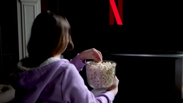 ティーン女の子座っています床にと時計Netflix上の大画面のテレビスクリーンセーバー遅い動きフラッシュ上の黒背景に赤青で若い女性のポップコーン女の子取りますそれと食べますそれ綴り — ストック動画
