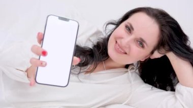 Beyaz ipek yatakta akıllı telefon kullanarak uzanan güzel esmer kadın manzarası. Yüksek kalite 4k görüntü