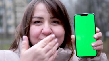 Olgun Asyalı iş kadını telefonunu gösteriyor. Kış-sonbahar kış gülüşü ile açık havada elinde yeşil krom bir telefonla öpüşen kıvırcık saçlı bir kadın. Harika bir neşe. 