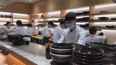 Maskeli ve eldivenli tanınmayan insanlar suşi restoranında Japon yemeği hazırlıyorlar. Beyaz önlüklü erkekler ve kadınlar steril giyiniyor. Herkes işini iyi yapıyor.