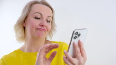 Cep telefonunu beyaz arka planda tutan kadın şüpheli ve şüpheci bir ifadeyle yana bakıyor..