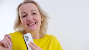 Neşeli beyaz kadın müzik dinliyor ve dans ediyor. Sarı saçlı, elinde telefon ve kulaklıklarla eğlendirici bir fotoğraf. Moda, teknoloji konsepti.