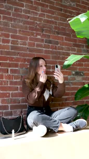 年轻美丽的嬉皮士快乐的女人坐在城市街道上 做化妆 青少年风格 平衡板 牛仔裤 运动鞋粉底 — 图库视频影像