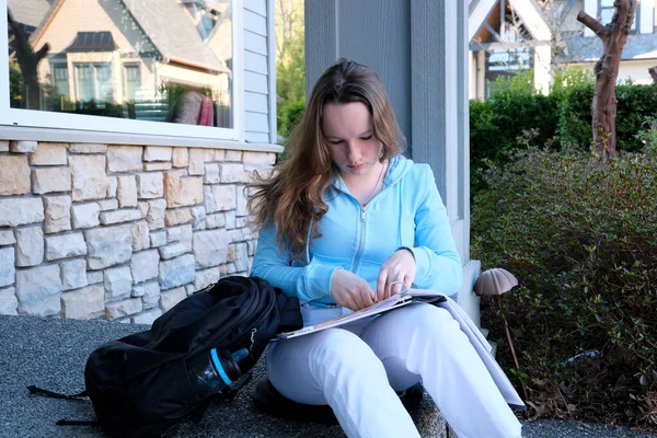 Stock image teenager girl does homework on porch of house she writes with pencil in large notebook notebook in folder next to backpack spring homework sit on threshold on porch woman writes draws solves problems