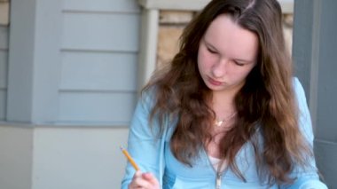 Genç kız ev verandasında ödev yapıyor. Kalemle büyük not defteriyle klasörde, sırt çantasının yanında bahar ödevi yapıyor. Verandadaki eşiğe oturuyor.