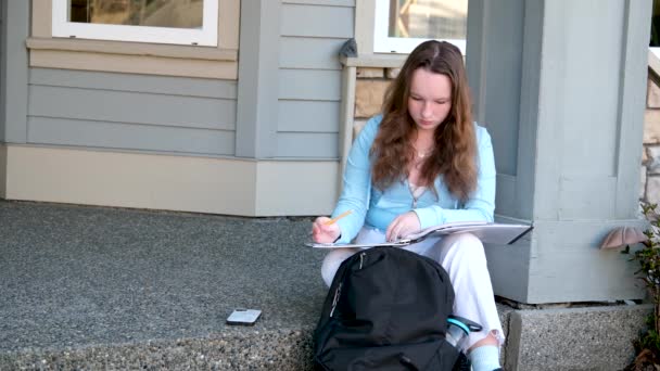 小女孩在门廊上做作业 她用铅笔在大笔记本的文件夹里写着 紧挨着背包 春天的作业坐在门廊上 — 图库视频影像