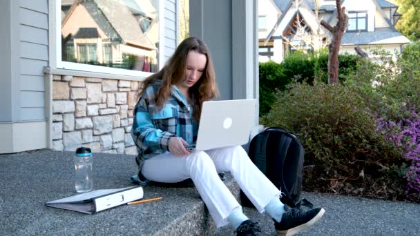 房间门廊上坐着一个带着笔记本电脑的少女 她在网上看新闻做家庭作业时哈哈哈大笑 — 图库视频影像