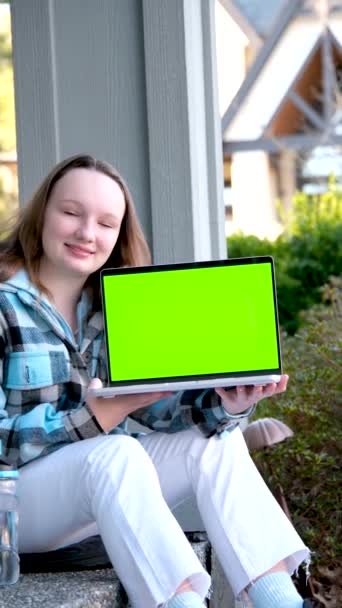 漂亮的少女手里拿着笔记本电脑 绿色荧幕着色键广告少女坐在门廊上检查蓝色夹克可爱的脸温柔的微笑看这里选这个可爱的产品满意 — 图库视频影像