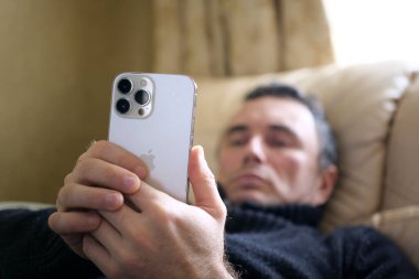 Yatakta uzanmış bir adam yeni telefon iPhone 13 Pro Max 'e bakıyor. Harika bir seçim. Sıradan biri.