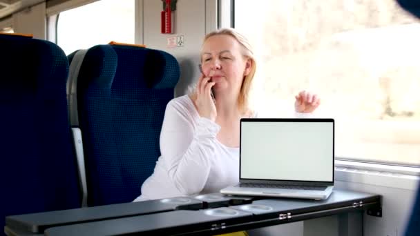 白い画面でノートパソコンの横の電車の中で女性が話している旅行トラフィックの女性は青い柔らかい席のテーブルに座っているビジネスクラスの美しい旅高品質 — ストック動画