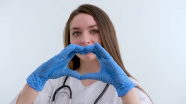 Gülümseyen genç bayan doktor kardiyolog beyaz bir ceket ve steteskop giyiyor. Kameraya bakarak kalp şeklini gösteriyor. Kardiyoloji sağlık hizmetleri aşk ve tıp yardım konsepti, portre