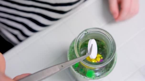 子供の手のための緑色の液体と金の輝き化学実験を持つボトルに大きな白い錠剤をドロップするためにトングを使用して白いテーブルの上に認識できない人々は見えません — ストック動画