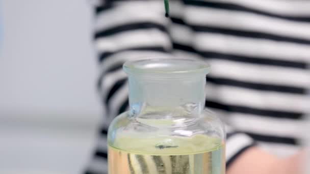 人の研究室実験の縞模様のブラウスを背景に青い緑色の液体滴を持つガラス瓶のクローズアップはゆっくりと瓶の底に落下 — ストック動画