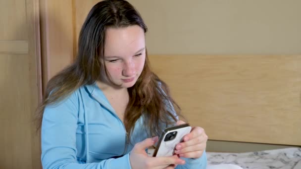 十代の女の子はベッドの上に座っています携帯電話を見て灰色の汚れと白いケース若い女性まっすぐ彼女の髪を取りたいですセルフィー通信オンライン — ストック動画
