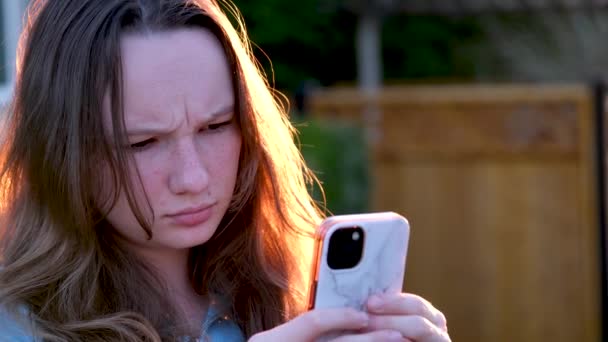 10代の女の子が街の夕日に眉をしかめっ面で何かに不満を持っている女の子が電話で新しいメッセージを見て不快な情報通信友人グループとソーシャルネットワーク — ストック動画