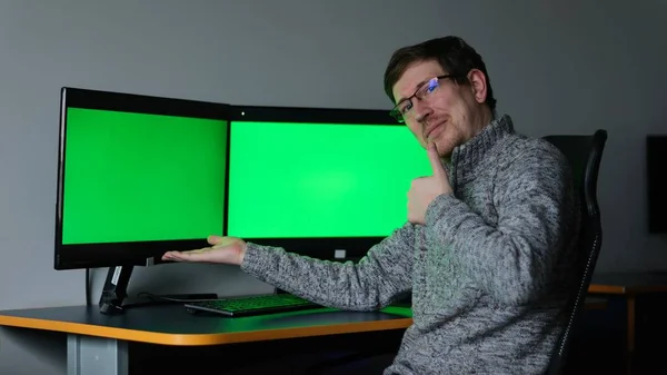 モニターの画面を反映したメガネの中年男性は 2つの画面でコンピュータの近くに座っています緑の背景クロマキー彼は手を示しており 私たちを見て自信を持って広告コンテンツグレーのセーター — ストック写真