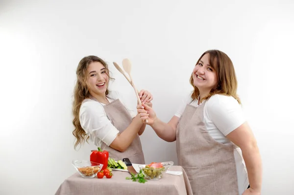 two women two friends mother and daughter adult and young fight on wooden spoons smile laugh in frame play have fun on the table knife vegetables nuts aprons of the same color cooking grocery store