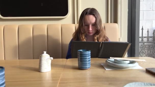 テーブル席の10代の女の子が紙のメニューを通して彼女の隣にお茶の青のジャケットのカップ緑の髪の明るい美しいレストランで慎重に異なる料理の選択を調べる食べ物の選択 — ストック動画