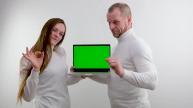 man and girl stand on white background in hands of laptop with green blank screen show gesture with little finger and forefinger goat magical sign protects from evil place for advertising satisfied