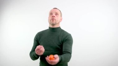 Bir adam mandalina hokkabazlığı yapıyor, hepsi yere düşüyor, onları komik yakalamaya çalışıyor ve beyaz arka planda eğiliyor, şaka yapıyor, eğleniyor adam mandalinayla hokkabazlık yapıyor. O çok kurnaz. Yüksek kalite