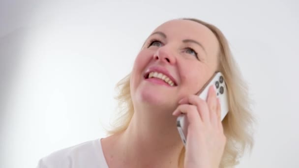 漂亮的金发女人在白色窗帘前听着手机里的一个电话 微笑着看着新闻的头和肩膀 高质量的4K镜头 — 图库视频影像