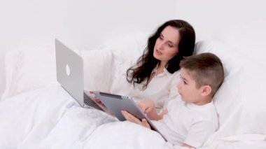 Anne ve oğlu beyaz battaniyenin altında yumuşak bir yatakta erkek tabletinin ellerinde sosyal ağlarla oynuyorlar. 4-6 yaşlarında kadın bilgisayarlı uzaktan kumanda konferansında serbest çalışıyor.