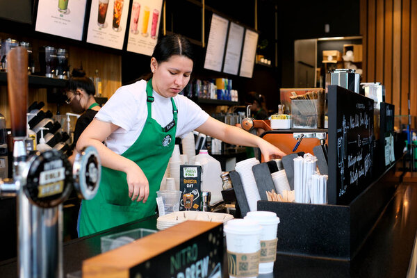 Starbucks in Canada in city of Vancouver preparing coffee of various drinks behind counter women in white t-shirt aprons and masked sellers in glasses of Asian-European ethnicity girl peeking