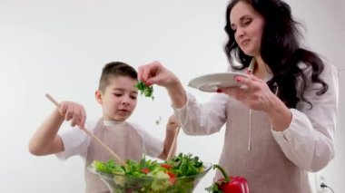 Güzel ev hanımı, küçük oğlunun taze marul yapraklarını parçalayıp açık modern mutfakta salata pişirmesine bakıyor.