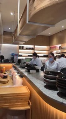 Restorandaki açık mutfak şefleri görülüyor. Garsonları hazırlıyorlar. Suşi siparişlerini eve götürüyorlar. Maskeleri takıyorlar.