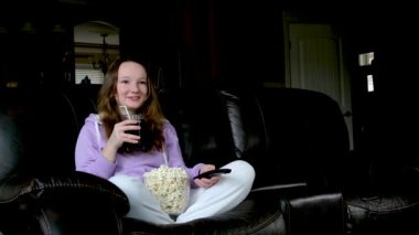 Şok olmuş küçük bir kız çocuğu komedi programı veya film içeriği izlerken evde tek başına rahat bir kanepede patlamış mısır yiyor. Küçük şirin okul çocuğu dinlenme zamanı..
