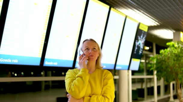 黄色の女性がテレビの前に立っている空港で問題を解決し 問題を解決するために会うことに同意するために飛行転送を待っている電話の深刻な人に話し合います — ストック動画