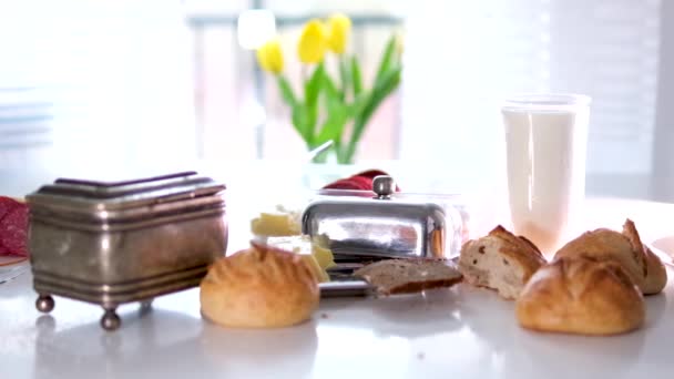 テーブルの上で実際の朝食壊れたパンパン粉バター砂糖ボウルミルクやケフィアガラス窓の太陽の下で3つの黄色のチューリップ白いテーブルリラックス実際の生活 — ストック動画