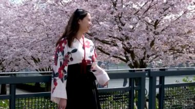 Yavaş çekim kızı, sakura ağacıyla neşeli bir şekilde dokunuyor. Uzun saçlı genç kadın bahar bahçesinin çiçeklenmesinden hoşlanıyor. Çiçek açan ağaçlarla Japon bahçesinde yürüyen kız.