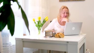 Kadın odasında oturmuş dizüstü bilgisayarla konuşuyor beyaz köşedeki yemek masasında kahve içiyor çiçeklerle çalışıyor serbest çalışıyor internette çalışıyor arkadaşlarıyla sohbet ediyor keyifli sabahlar geçiriyor.