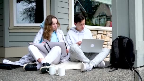 10代の若者がノートパソコンとタブレットを使って家庭からオンライン教育を受けます隔離の概念はあなたの耳の中のポーチのささやきの外に座っています大きな白いフォルダに宿題ノートパソコンをやって楽しい笑いがあります — ストック動画