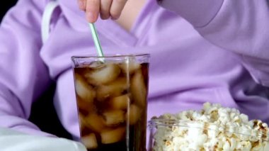 Yakın plan bir bardak Coca-Cola ve cam tabağın yanında patlamış mısırlı, mor kazaklı kız pipetli, tanınmayan insanlar hafta sonu film izleyip dinleniyorlar.
