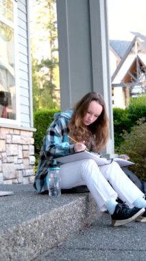 Bir kız, büyük bir defterde kalemle çiziyor. Defterler, köy evi şişesi, beyaz kot pantolon, siyah spor ayakkabı, damalı mavi ceket, uzun kahverengi saçlı genç kız.