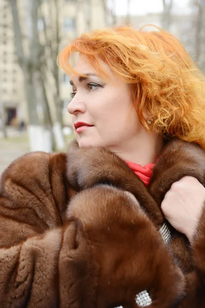 Руда жінка в білому в'язаному капелюсі і одягнена в теплий з капюшоном повсякденний одяг для паркету, дівчина на парковці, пішохідний перехід і автомобіль на задньому плані, дівчина зігрівається — стокове фото