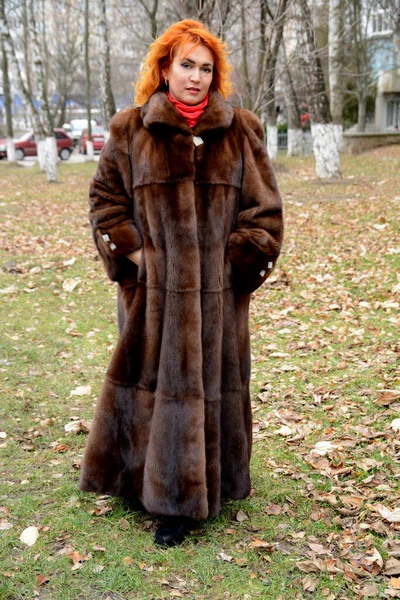 red-haired beautiful middle-aged woman in long fur coat in the park forest on the street walks looks into the frame red scarf bright brown mink coat