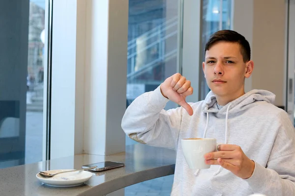 若いハンサムな男がクロスを感じ親指を下げてコーヒーカップを持ってる 高品質の写真 — ストック写真