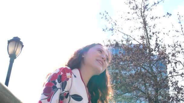 温哥华在摩天大楼的背景下 从下面闪烁着美丽的年轻姑娘的手电筒 背景是春树的微笑 头发被风中的亚洲女人吹散在绣花衬衫上 — 图库视频影像