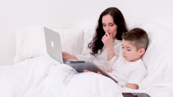躺在床上 躺在床上 母亲咳嗽着 躺在自己四五年大的儿子旁边 手里拿着平板笔记本电脑 等待着康复 — 图库视频影像