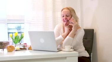 Bilgisayarda çalışan orta yaşlı bir kadın telefonda gülüyor çay içiyor kahve içiyor sabah kahvaltısı güneş ışığı pencerenin önünde çiçekler açıyor hoş bir sohbet.