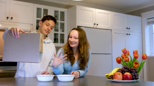 幸せな男の子と女の子は家で朝の健康的なシリアル朝食のためのシリアルボックスの外に焼きチョコレートボールを注ぐ紙箱から白いプレートにコーンフレークを注ぐ — ストック動画